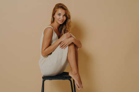 Photo of pleased young female model wears dress has slender legs crumpled up sits on chair against beige studio wall smiles gently blank space to place your information. Women natural beautyの写真素材