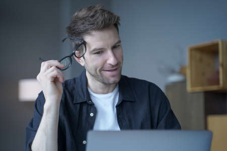 Attractive positive businessman in casual wear took off his glasses, remotely working from home office on new company project, focused European entrepreneur looking at laptop screen with head tiltedの写真素材