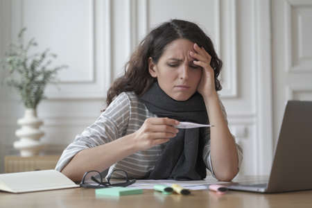 Unhealthy italian woman wrapped in warm knitted scarf looking anxiously on digital thermometer in...
