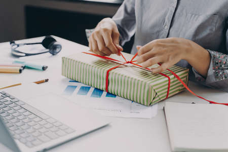 Cropped image of young businesswoman packing and decorating with ribbon Xmas gift while sitting at her workplace with laptop and stationary, preparing presents for colleagues at work at Christmas timeの写真素材