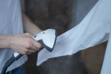 Cropped photo of man using steamer neatly ironing white shirt while holding long sleeve. Businessman tidies up formal outfit before important company meeting in officeの写真素材