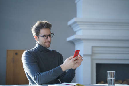 Young handsome german man in glasses sitting at table and using mobile phone, smiling austrian...