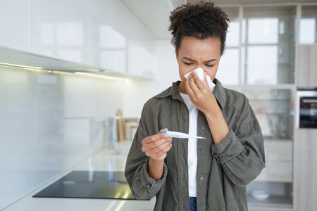 At home, a sick mixed race girl blows her nose and checks her temperature with an electronic...