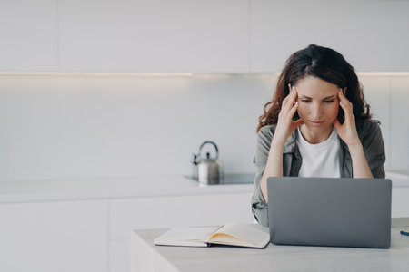 Stressed freelance worker solves laptop problems, suffers from migraine at home. Overworked woman touches temples, has headache during computer work.の写真素材