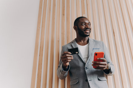 Excited African-American businessman shopping online, using smartphone and credit card, ordering food and making a long-awaited purchase via e-banking.の写真素材