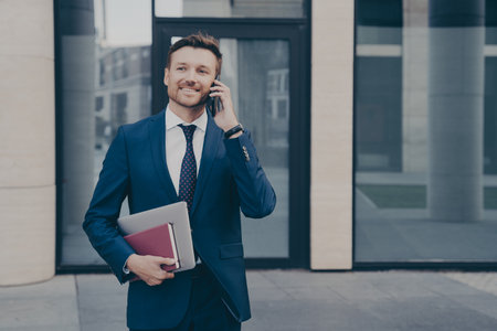 Confident owner in formal wear, giving directions on phone to employee, holding notebook and laptop, blurred background.の写真素材