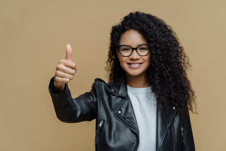 Pleased woman with curly hair, thumbs up, rates excellent product, recommends promo offer. Glasses, leather jacket. Isolated beige background, approves best deal.の写真素材
