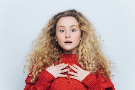 Curly-haired woman in red sweater, stunned gaze at camera, isolated on white background. Scared yet captivating.の写真素材