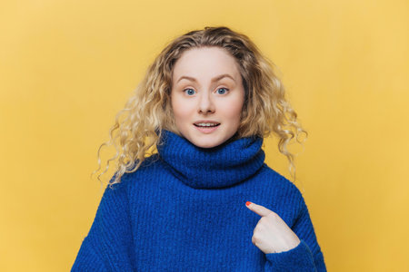 Surprised female model with curly hair points to empty space on her new winter sweater, perfect for your ad or design, on yellow background.の写真素材