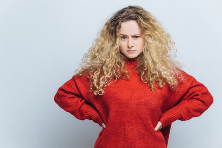 Angry woman with curly hair, hands on waist, loose sweater. Displayed expression. Isolated on white background. People, negative emotions.の写真素材