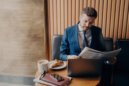 Focused entrepreneur in formal wear reads financial chronicle, coffee break with cappuccino, croissant, laptop open.の写真素材