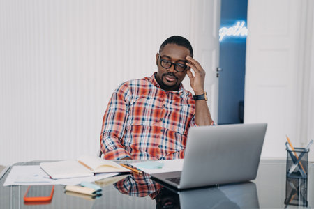 Overworked black employee: headache from long laptop work at desk. Tired businessman closes eyes,...