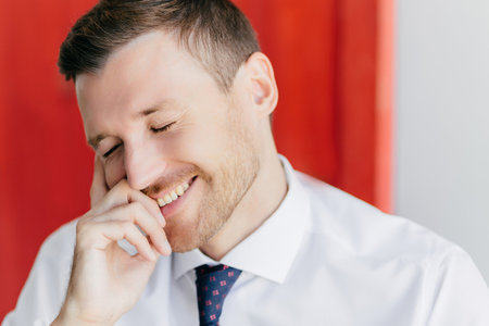 Joyful male in white shirt giggles at funny joke, closes eyes with pleasure. Receive pleasant news. Poses against red wall. positive emotions concept.の写真素材