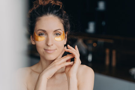 Confident woman using collagen eye patches. Happy Hispanic girl post-bath, spa therapy. Skincare, cosmetics, anti-aging beauty.の写真素材