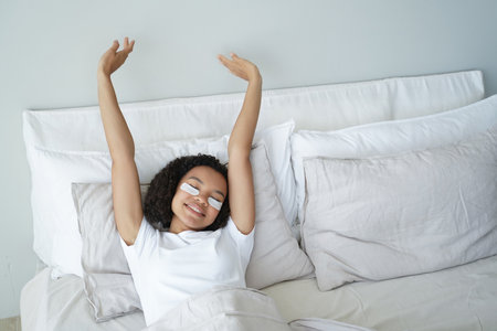 Happy girl stretches in bed with eye patches on face. Morning beauty routine.の写真素材