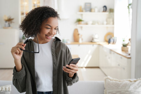 Smiling mixed-race girl with afro hairstyle using smartphone, shopping online, and chatting on social networks. Modern teen reading good news at home.の写真素材