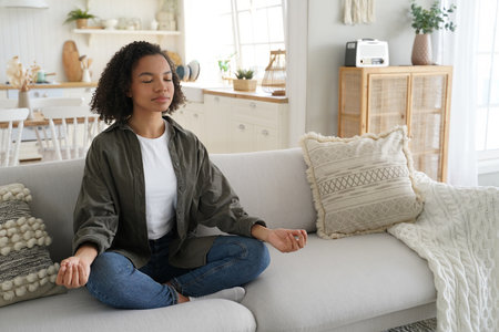 Serene mixed-race girl meditates at home, in lotus pose with mudra gesture on the sofa. Afro-haired, calm, practicing yoga. Healthy lifestyle, meditation.の写真素材