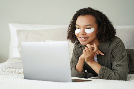 Mixed race girl relaxes with eye patches, watches series on laptop. Skincare ad.の写真素材