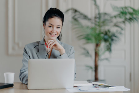 Cheerful professional woman searches laptop update, removes glasses in formal office attire, working remotely on blog ideas.の写真素材