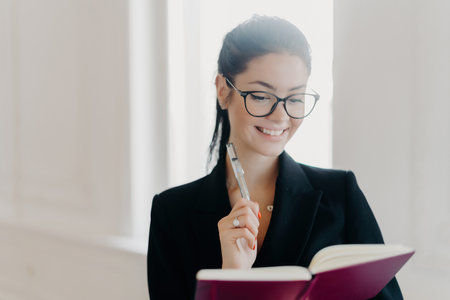 Glad prosperous woman CEO in formal clothes and transparent eyeglasses, smiling as she writes in notepad, planning work schedule and noting startup ideas indoors.の写真素材