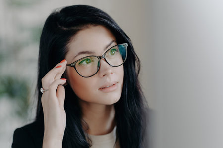 Serious brunette with spectacles, focused on screen, browsing web, creating projects indoors, office setting.の写真素材