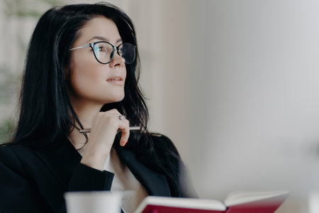 Thoughtful woman notes ideas, plans, and goals in organizer with glasses on, deep in thought.の写真素材