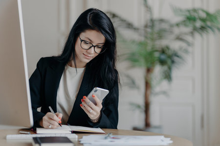 Focused woman with dark hair searches online, takes notes, at desk surrounded by papers.の写真素材