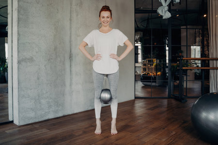 Joyful redhead woman in white tee and gray leggings does Pilates in a fitness studio. Holds silver...