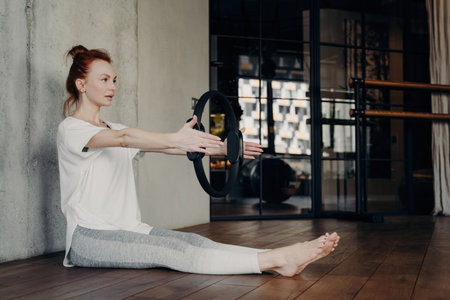 Focused sporty woman in white t-shirt and gray leggings sits in fitness studio, doing arm exercises with Pilates ring. Legs stretched.の写真素材