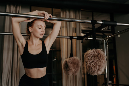 Exhausted redhead sportswoman in black top takes a rest, leaning on the Cadillac reformer after Pilates exercises. Enjoying her workout time in the studio with special indoor sports equipment.の素材