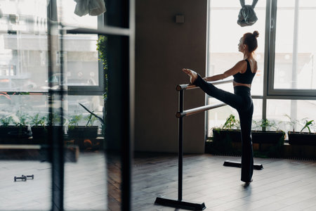 Slim red-haired ballerina stands in split position beside ballet barre in gym, light window background. Indoor sport and stretching showcased beautifully.の写真素材
