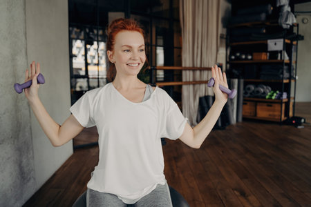 Happy European redhead woman in active wear sits on silver fitball, raising arms with dumbbells in fitness studio. Positive workout vibes!の写真素材