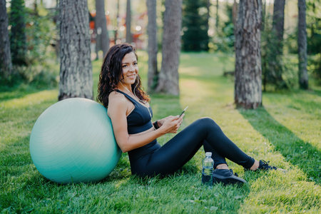 Satisfied brunette woman sits against big fitness ball, using her phone for music, on green grass in a sunny forest. Sporting concept, wearing sports clothes.の素材