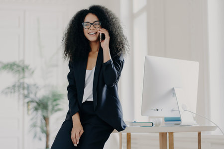 Satisfied dark-skinned businesswoman on break, leaning on white table with computer in her office, talking on her cellphone, wearing glasses and an elegant suit, consulting with a partner.の素材