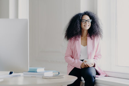 Joyful female entrepreneur, Afro hair, rosy jacket, sips coffee by window in spacious office with modern setup.の素材