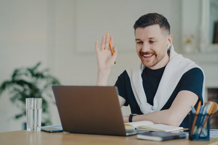 Cheerful freelancer/businessman in earphones waves, greets colleagues/clients in video conference from home office.の写真素材