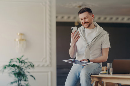 Bearded freelancer in coworking space, video calling on smartphone with wireless earphones, free high-speed internet, having an online conference with coworkers.の写真素材