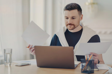 Busy freelancer in casual wear analyzes project results, works remotely from home office, multitasking with computer and paperwork. Embracing freelance and business concepts.の写真素材