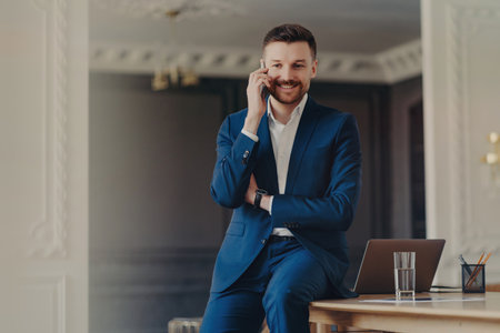 Handsome bearded man in blue suit, satisfied with good news, talking on the phone, posing at desk with modern laptop. mobile conversation.の写真素材