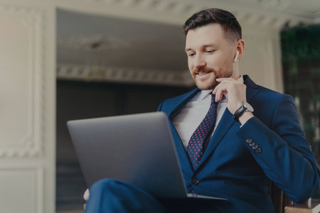 Confident businessman in formal suit, wearing wireless earphones, looking at laptop screen during online business meeting. Discussing project with colleagues in a luxury hotel or office.の写真素材