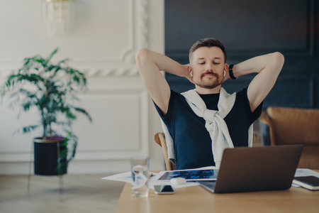 Relaxed man freelancer or entrepreneur, wireless earphones, hands behind head, watching webinar on laptop, working from home.の写真素材