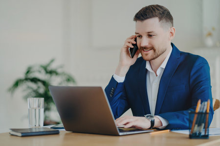 Successful young entrepreneur in formal suit smiles, talks on phone, discusses business with partner in modern office.の写真素材