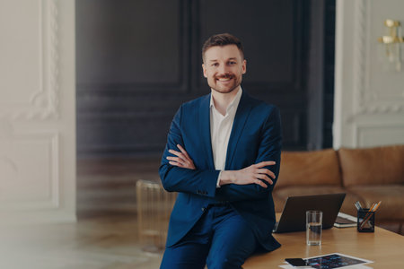 Satisfied bearded businessman in formal attire, stands confidently in office, arms folded, smiling pleasantly, content with work results.の写真素材