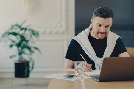 Focused freelancer in casual wear notes on laptop at home workspace. Manager gathers feedback or prepares a project report.の写真素材