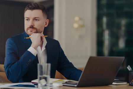 Serious male entrepreneur in blue suit, stylish office, thinking about business challenges, holding pencil, focused.の写真素材