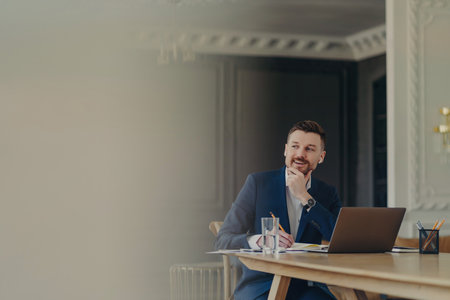 Successful bearded businessman in dark blue suit, writing business plan in notepad at stylish office/luxury apartment, with laptop nearby, looking happy.の写真素材