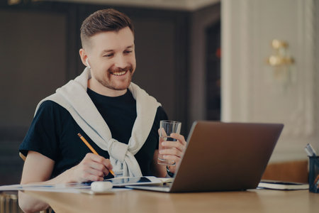 Happy man freelancer/entrepreneur, casual wear, laptop surfing, holding water glass, working online from home. freelance concept.の写真素材