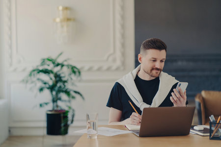 Casual businessman multitasks with smartphone, notes important contacts, and watches business webinar on social media. Freelancer works on laptop from home office.の写真素材