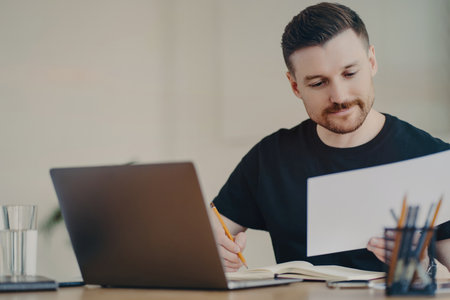 Bearded expert edits reports on laptop, focused at home office, casual in black t-shirt.の写真素材