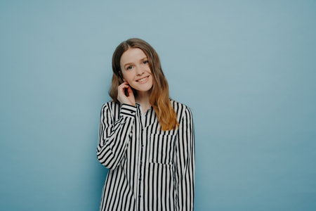 Tender cute European teen girl in striped white shirt, tilting head with hair behind ear, expressing shyness, posing in studio against light blue background.の写真素材